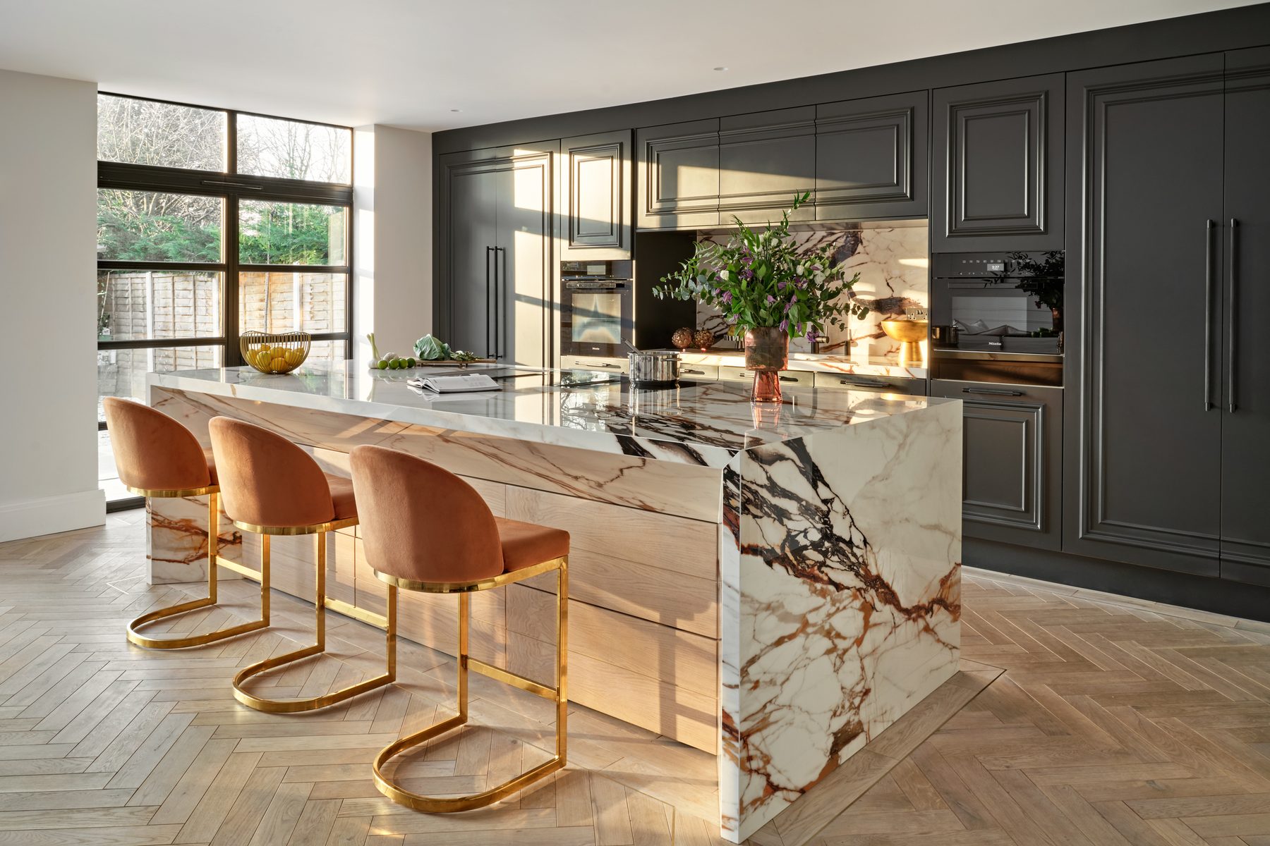 A finished Sheraton kitchen with dark panelled cabinetry, a dramatically veined waterfall island and blush bar stools
