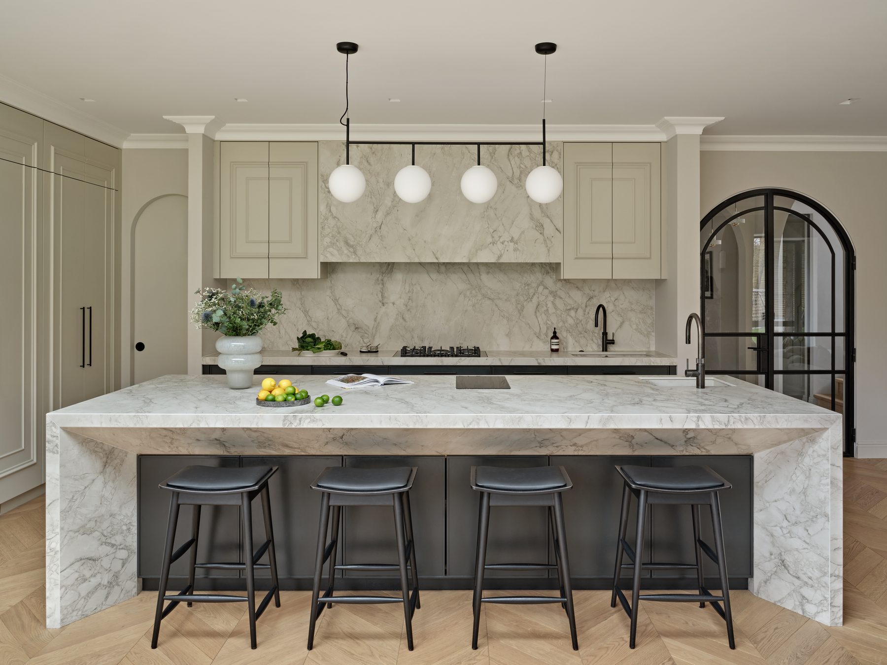 A finished Sheraton kitchen in soft stone tones with a large veined waterfall island, globe pendant lights and black bar stools