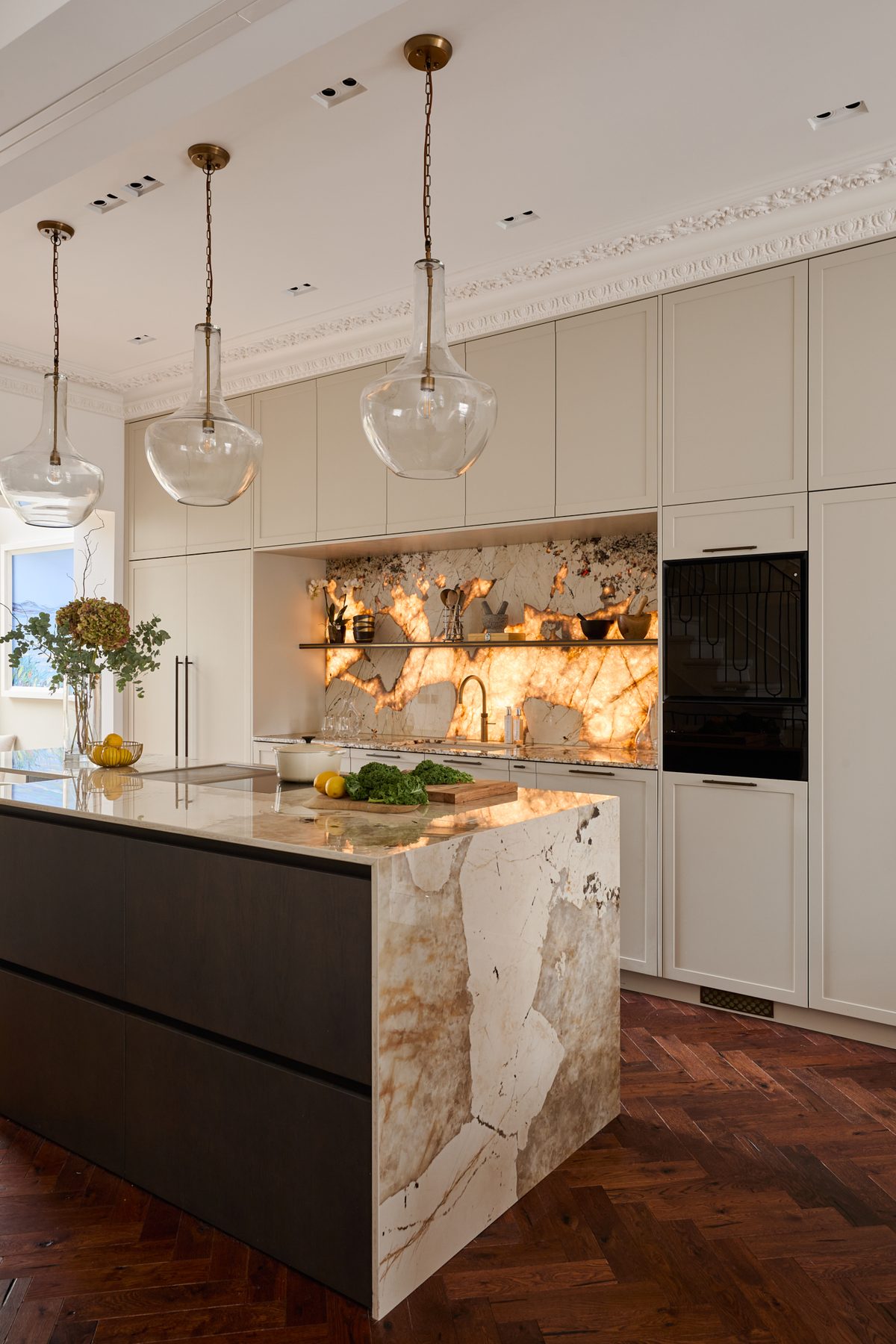 A finished Sheraton kitchen with cream cabinetry, a dark island with a veined waterfall end and a lit shelf niche